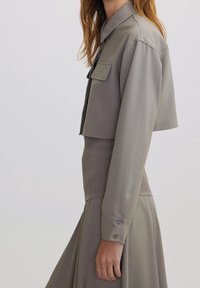 Femme portant une veste cropped taupe sur une jupe côtelée taille haute, montrant le profil latéral du cou jusqu'au milieu de la cuisse sur un fond uni.