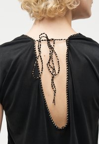 Ξανθό άτομο με σγουρά μαλλιά που φοράει μαύρο τοπ με βαθιά ανοιχτή πλάτη, δεμένο στο λαιμό με πλεγμένα μαύρα και μπεζ κορδόνια.