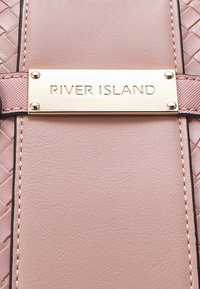 Jasnoróżowa torebka z plecionej tekstury, gładkiego materiału i wypolerowanej złotej plakietki z napisem "RIVER ISLAND" na środku.