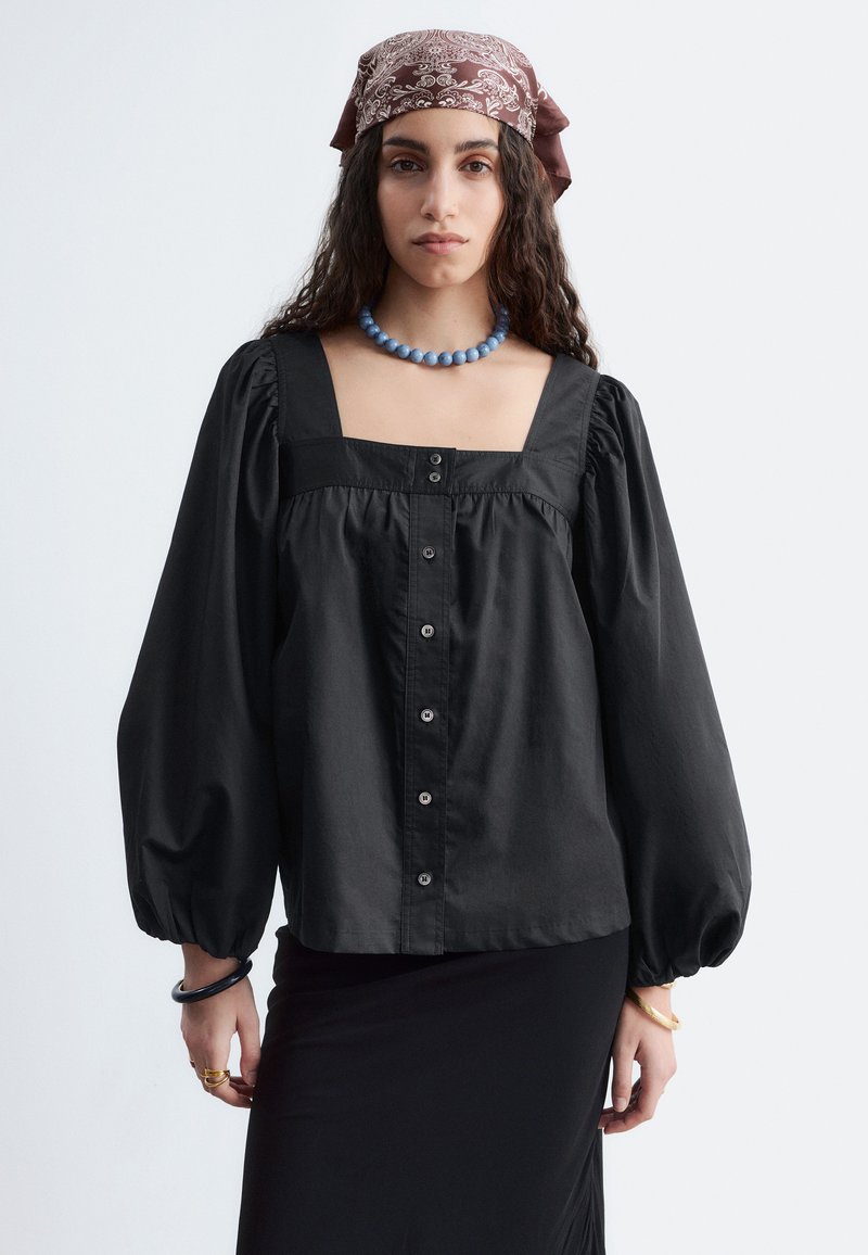 Femme aux longs cheveux bouclés portant un foulard à motifs, un collier de perles bleu, un chemisier noir ample boutonné avec des manches bouffantes et une jupe noire.