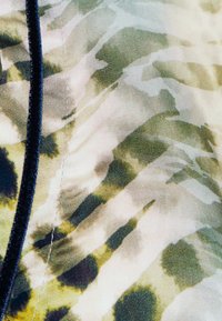 Vihreän ja kerman sävyinen teksturoitu kangas, jossa on abstrakteja raitoja ja läiskiä. Reunus tummansinertävällä nauhalla.