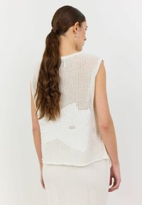 Vit stickad topp med blommönster, lös passform och utan ärmar, med en öppen stickad struktur och sidoknutar.