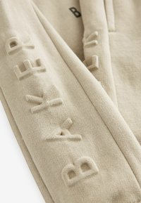 Suéter beige con letras texturizadas y en relieve. Presenta un diseño sencillo y material de punto suave, adecuado para un uso casual.