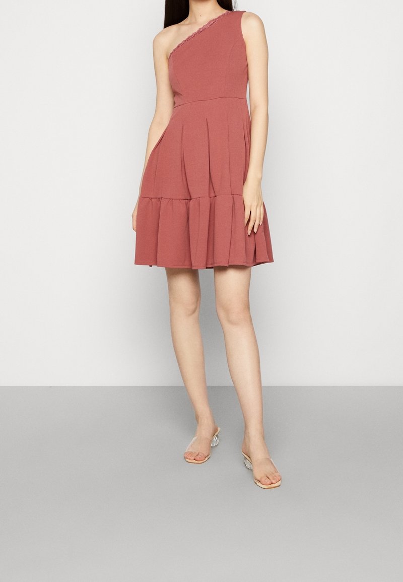 Femme portant une robe rose poudré une épaule sans manches avec un ourlet volanté et des talons transparents ouverts à l’avant, debout devant un fond uni.