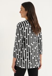 Vrouw met lang bruin haar draagt een zwart-wit geometrisch patroon met lange mouwen en zwarte broek, staat zijwaarts op een eentonige achtergrond.