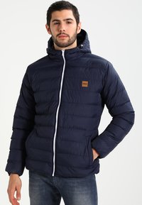 Navy blue puffer jacket with a hood, featuring a zip front, white zipper, and a small brown logo patch on the chest. Smooth texture.