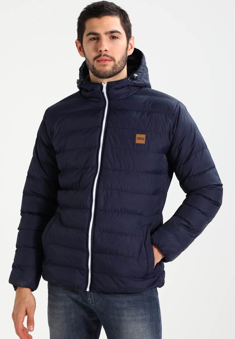 Navy blue puffer jacket with a hood, featuring a zip front, white zipper, and a small brown logo patch on the chest. Smooth texture.