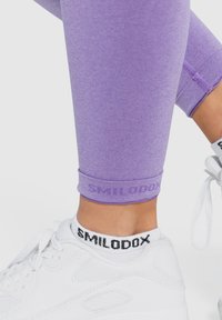 Lila Sportleggings mit glatter Textur, ausgestattet mit dem „SMILODOX“-Logo am Saum. Kombiniert mit weißen Turnschuhen und Socken mit schwarzen Akzenten.