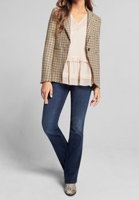 Femme portant un blazer pied-de-poule moutarde, un chemisier crème à volants, un jean bootcut bleu foncé et des bottines en peau de serpent, debout dans un espace neutre.
