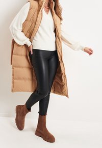 Tan padded vest over a white sweater, black leather leggings, and brown suede ankle boots with chunky soles. Simple design, soft textures.