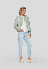 Texturiertes, hellblaues, gewebtes Jackett mit goldenen Knöpfen, über einem weißen T-Shirt mit einem dezenten Design getragen, kombiniert mit ausgebleichten hellen Jeans und beigen Slippern.