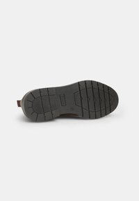 Suela de zapato de ante marrón con una plantilla de goma negra texturizada. Presenta un dibujo en la suela para mayor tracción, con una pequeña asa.