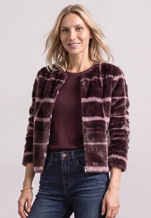 Blonde vrouw draagt een paars-roze gestreepte nepbontjas, een bordeaux top en blauwe spijkerbroek, glimlachend tegen een effen achtergrond.