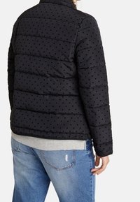 Black puffer jacket with dotted texture, featuring quilted design, high collar, and visible stitching. Worn over a gray top and blue jeans.
