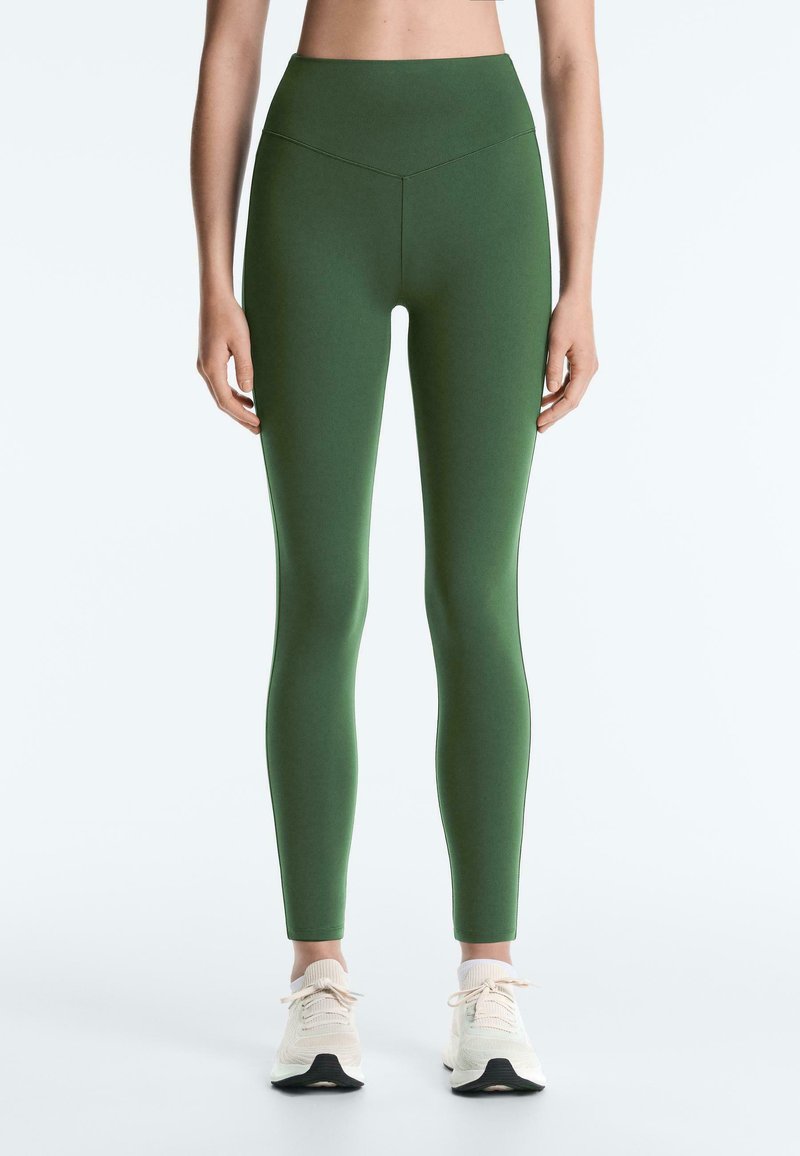 Femme portant un legging vert taille haute et des baskets blanches, debout avec les bras détendus le long du corps devant un fond uni.