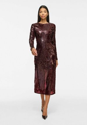 Femme portant une robe midi à manches longues rouge foncé à sequins et des escarpins noirs pointus, marchant vers l’avant sur un fond blanc uni.