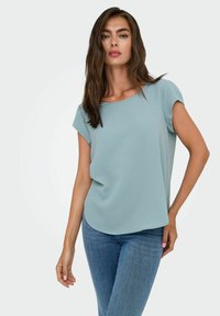 Frau mit langen braunen Haaren, die eine hellblaue Bluse mit kurzen Ärmeln und blaue Jeans trägt, posiert mit einer Hand auf der Hüfte.