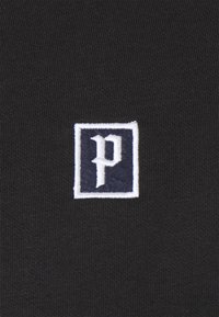 Schwarzer Stoff mit einem rechteckigen gestickten Aufnäher mit einem weißen Rand und einem stilisierten weißen Buchstaben "p" in einem marineblauen Quadrat.