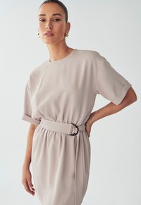Knie-lange, licht beige jurk met een ronde hals, korte mouwen, een aansluitende taille en een riem met gespsluiting. Glad materiaal met een zachte drape.
