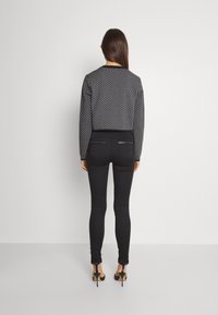 Svart mönstrad tröja med geometrisk design, kombinerad med svarta slim fit-jeans och högklackade sandaler. Sett bakifrån mot en enfärgad bakgrund.