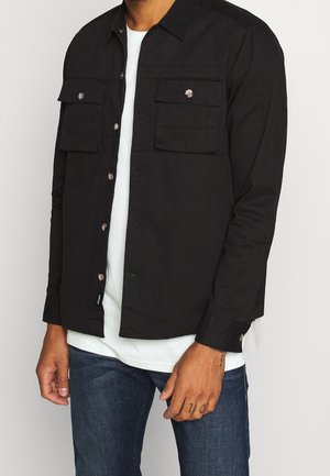 Mand iført sort skjorte med knapper og to brystlommer, en hvid undertrøje og blå jeans mod en ensfarvet baggrund.