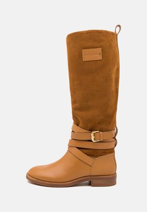 Botte haute en cuir et daim couleur fauve avec un bout arrondi, deux brides croisées avec une boucle dorée, et une texture douce.