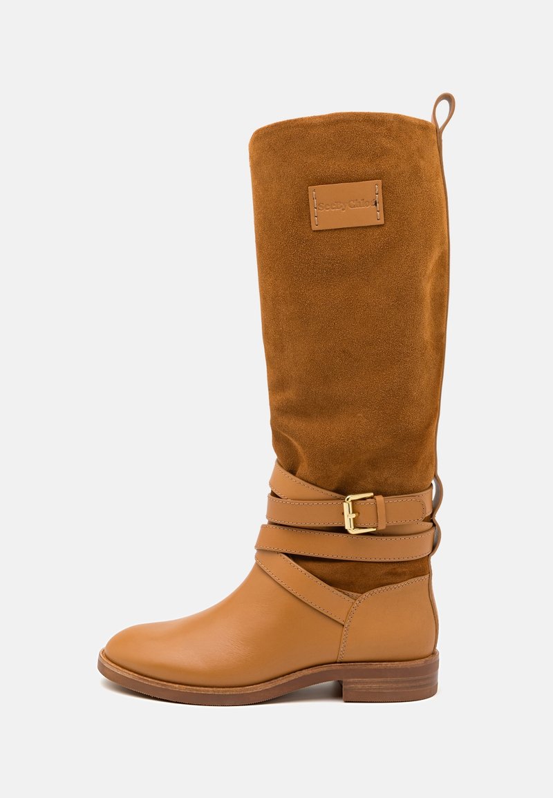 Botte haute en cuir et daim couleur fauve avec un bout arrondi, deux brides croisées avec une boucle dorée, et une texture douce.