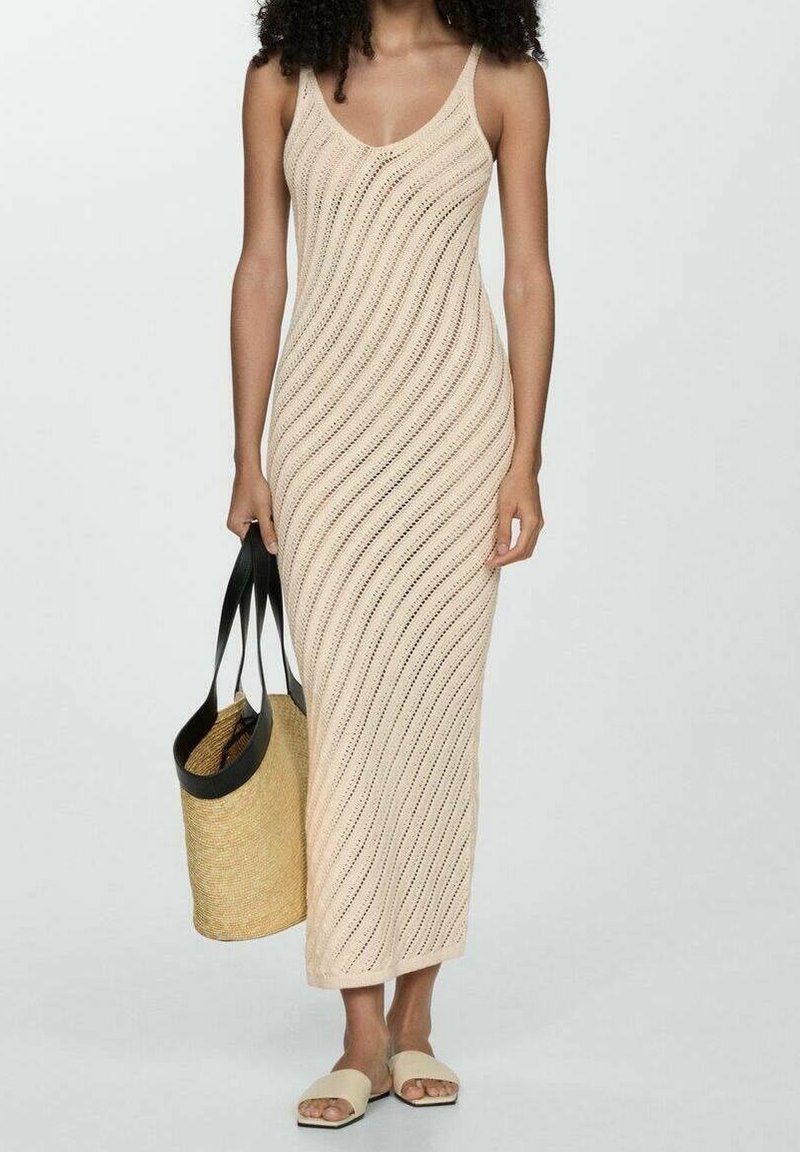 Femme portant une robe maxi en tricot beige sans manches avec des motifs diagonaux, tenant un sac cabas en paille avec des poignées noires et portant des sandales slides beiges.