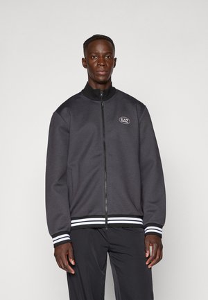 Gray jacket with a high collar, front zipper, and white striped accents at the cuffs and hem, featuring an embroidered logo on the chest.