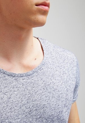 Untere Gesichtspartie und Hals eines jungen Menschen, der ein hellgraues, gesprenkeltes Rundhals-T-Shirt vor einem einfarbigen Hintergrund trägt.