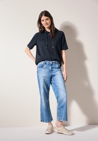 Top negro de manga corta con cordón, jeans de pierna ancha azul claro y zapatillas beiges con cordones. Texturas suaves y ajuste relajado.