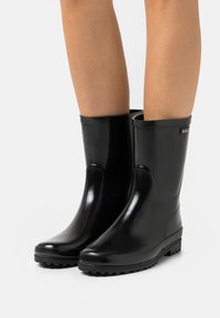 Bottes en caoutchouc noires imperméables avec une texture lisse et brillante, un bout arrondi et un petit talon. Présente un logo en relief sur le côté.