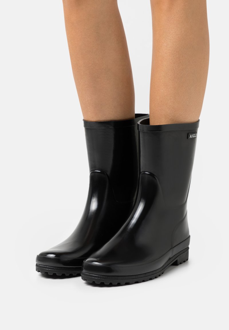 Bottes en caoutchouc noires imperméables avec une texture lisse et brillante, un bout arrondi et un petit talon. Présente un logo en relief sur le côté.