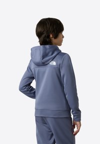 Μπλε φούτερ με κουκούλα, με λευκό λογότυπο στο πάνω μέρος της πλάτης. Λεία υφή και εφαρμοστή σχεδίαση, με μακριά μανίκια και κομψή γραμμή.