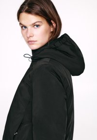 Crna jakna s kapuljačom od mekog materijala, s kapuljačom koja se može stezati. Ima jednostavan, prianjajući kroj i minimalistički dizajn bez vidljivih kopči.