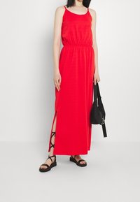 Robe maxi rouge avec une taille cintrée, des fentes latérales et des bretelles spaghetti. Associée à des sandales noires et un petit sac noir. Texture de tissu lisse.