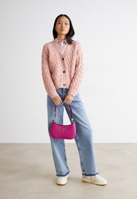Strikket lyserød cardigan med et vævet mønster, brune knapper, parret med lyseblå jeans og hvide sneakers, holder en lyserød håndtaske.