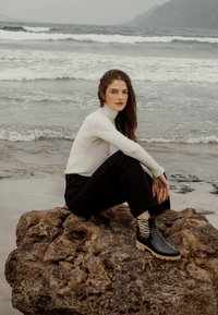 Botas negras y brillantes hasta el tobillo con una suela resistente, combinadas con un cuello de tortuga blanco y jeans negros, sentadas sobre una roca junto a la orilla.