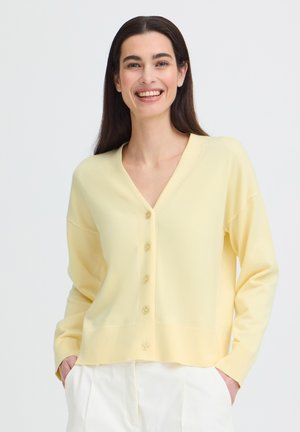 Mosolygós nő hosszú sötét hajjal, világos sárga gombos kardigánt és fehér nadrágot visel, kezét zsebében tartva.