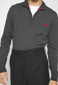 Pullover grigio con zip e un accento rosso al colletto, realizzato in tessuto a maglia. Abbinato a pantaloni neri su misura.