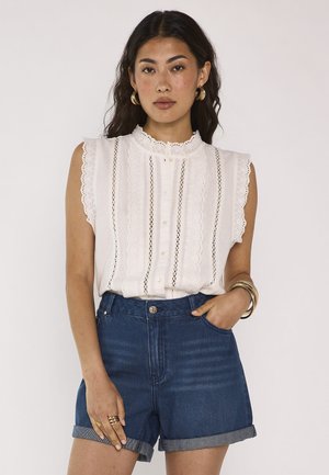 Femme aux longs cheveux foncés porte un chemisier blanc en dentelle sans manches, un short en jean taille haute roulé, des créoles dorées et des bracelets.