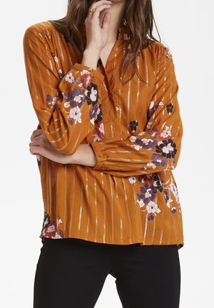 Mujer vistiendo una blusa naranja óxido con rayas metálicas verticales y patrones florales, combinada con pantalones negros, con la mano cerca de la cara y el brazo cruzado.