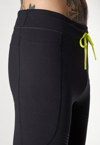 Schwarze Sportleggings aus dehnbarem Material, mit einem hellgelben Kordelzugverschluss und perforierten Details an den Seiten.