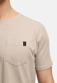 Beige Baumwoll-T-Shirt mit kurzen Ärmeln, rundem Ausschnitt und einer Brusttasche mit schwarzem Etikett. Weiche Textur, lässige Passform.