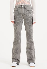 Jeans a zampa d'elefante grigi con intricati motivi neri ricamati. Design standard a cinque tasche con chiusura a bottone e branding discreto sul davanti.