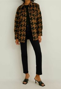 Giacca camicia a pince con motivo a scacchi marrone e verde, con chiusura a bottoni, due tasche sul petto e vestibilità rilassata, abbinata a jeans skinny neri e tacchi aperti.