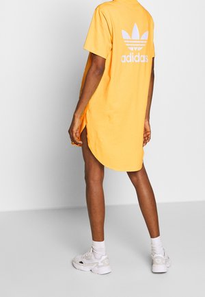 Osoba ubrana w żółtą sukienkę T-shirt Adidas z białym logo na plecach, białe skarpetki i białe trampki, stojąca na tle jednolitego szarego tła.