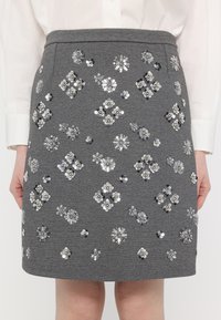 Jupe grise avec des motifs floraux en sequins argentés portée avec une chemise blanche à manches longues, les mains reposant à côté.