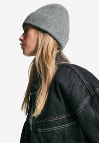 Bonnet en tricot côtelé gris porté avec une veste en denim noire présentant des coutures orange contrastantes. La veste a une coupe structurée, légèrement surdimensionnée.