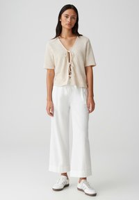 Knit, beige short-sleeve cardigan with a V-neck and tie closure at the front, paired with white wide-leg trousers and white sneakers.